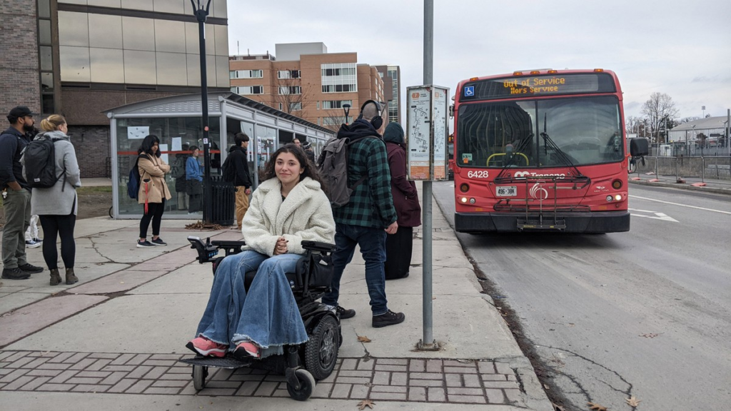 Jessica Ursitti at Carleton University's bus stop. People are scattered around, waiting for a bus.