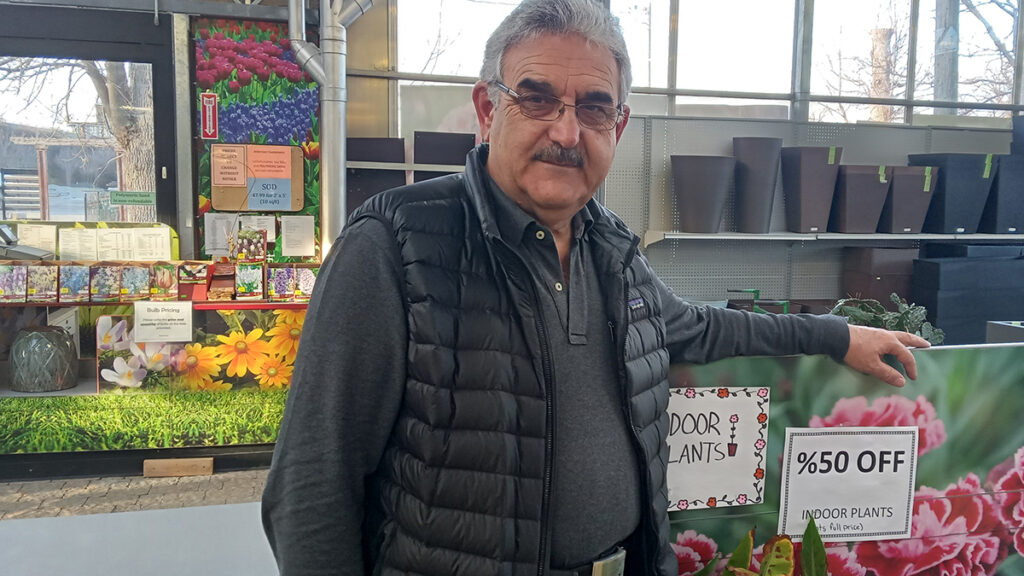 A tall light skinned man standing beside and behind some indoor flowers. He is wearing a black long sleeve and a winter jacket.
