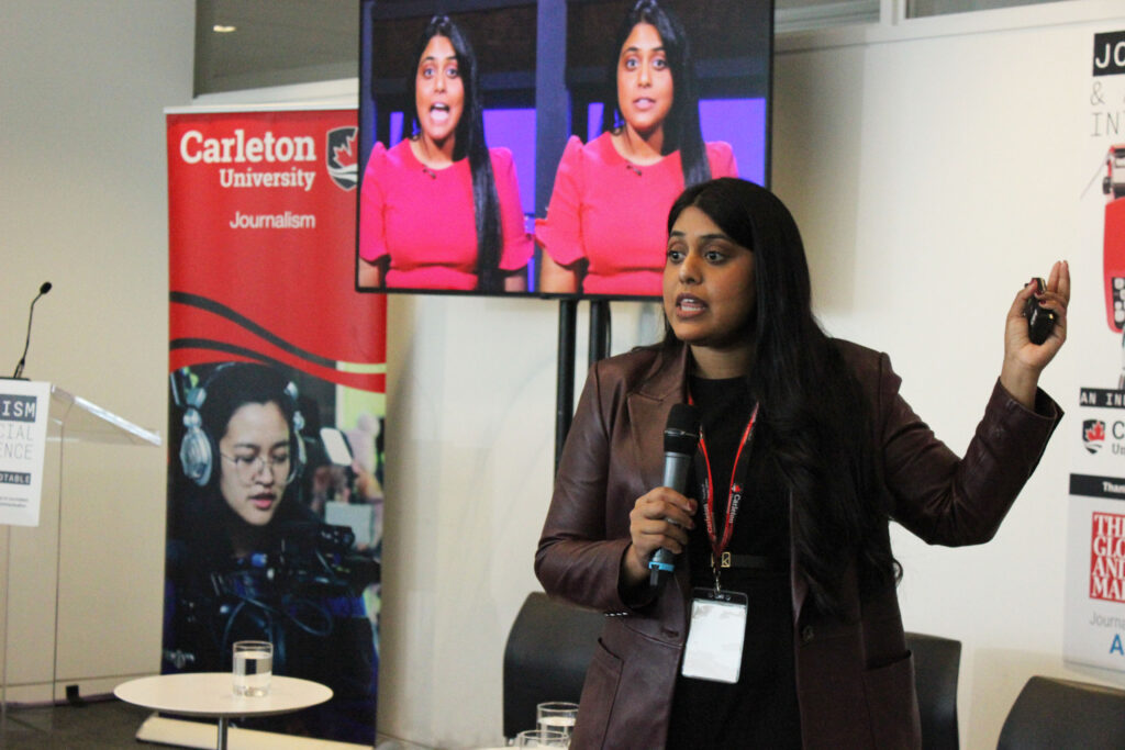 Nikita Roy, a speaker at the Carleton University Journalism event, is seen presenting with a microphone in hand. She wears a brown jacket and a lanyard with an ID badge, gesturing as she addresses the audience. Behind her, a large screen displays two images of her in a red dress. A Carleton University Journalism banner and a podium are also visible in the background.
