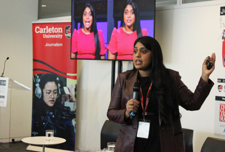 Nikita Roy, a speaker at the Carleton University Journalism event, is seen presenting with a microphone in hand. She wears a brown jacket and a lanyard with an ID badge, gesturing as she addresses the audience. Behind her, a large screen displays two images of her in a red dress. A Carleton University Journalism banner and a podium are also visible in the background.