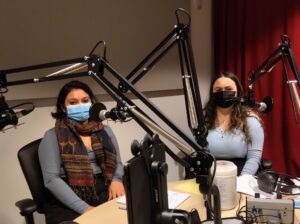 Two hosts in studio amid mics, wearing masks.