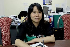 Dinh Thi Thuy sits in her office. She looks directly into the camera, her face is blank. In front of her rests a pad of paper and her cellphone. She is holding a pen in her hand, that's pointing straight up. She sits in a metal framed chair that has a thick red fabric upholstered to the back. Behind her sits a desk that is completely covered in stacks of white paper. There doesn't appear to be any room to work.