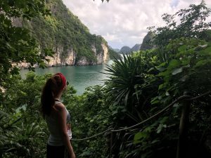 The young female author, Jordan Steinhauer, has her back facing the camera. She is surrounded by deep green bushes and trees. In front of her is a roped pathway. Peeking out between the leaves is bright turquoise water. Sitting within the water are a series of stone mountain peaks with green trees resting on top of the light stone. White clouds are visible between the mountains.