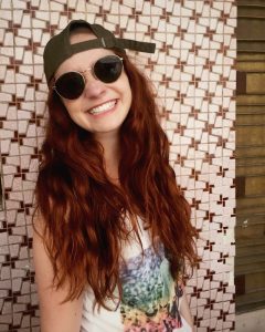 The young female author, Jordan Steinhauer, is smiling widely. Her long red hair is large and wavy from the Vietnam humidity. She is wearing round wired sunglasses and a baseball cap that has been turned backwards. She is wearing a white tank top that has a rainbow colored image on the front. She stands in front of a patterned tiled wall.