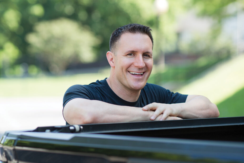 Trey Tucker leans against a car and smiles at the camera 