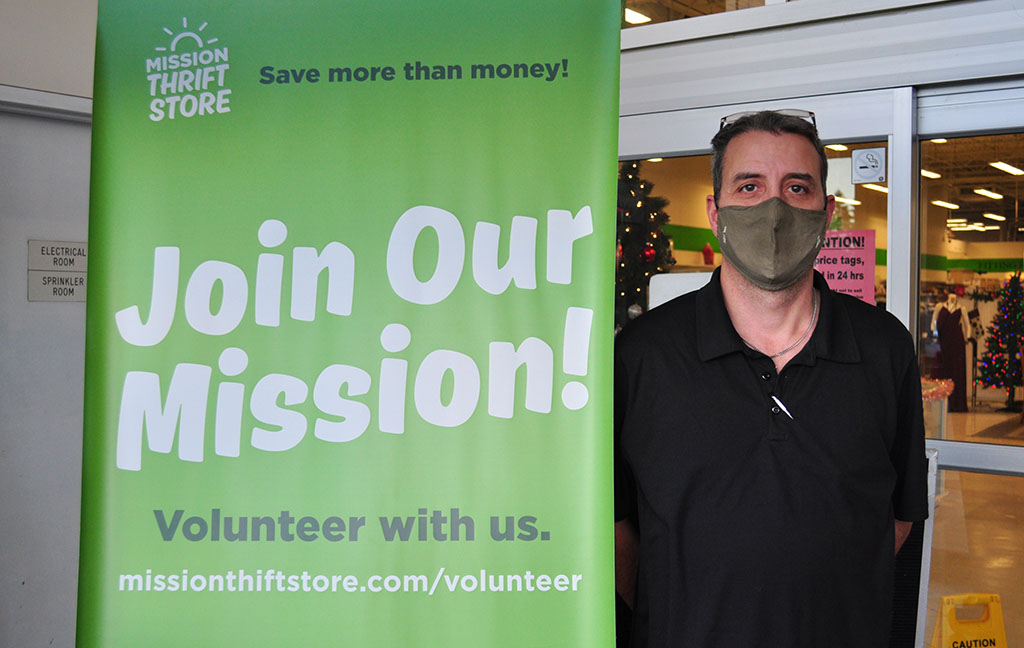 a man standing beside a green sign by Mission Thrift Store that reads "Join Our Mission!"
